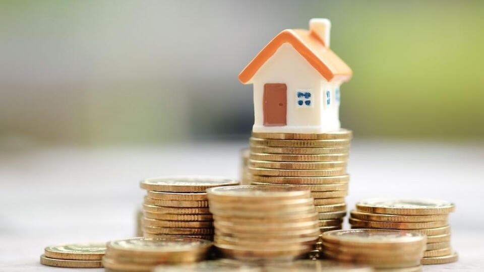 Miniatuurhuisje met oranje dak op stapels gouden munten tegen een wazige achtergrond.