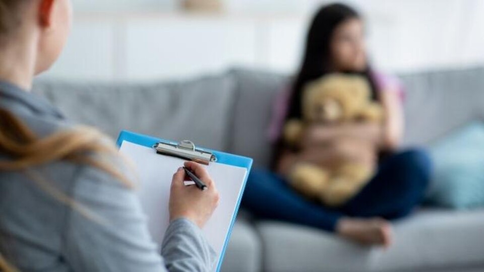 Hulpverlener schrijft op clipboard terwijl kind met teddybeer op bank zit.