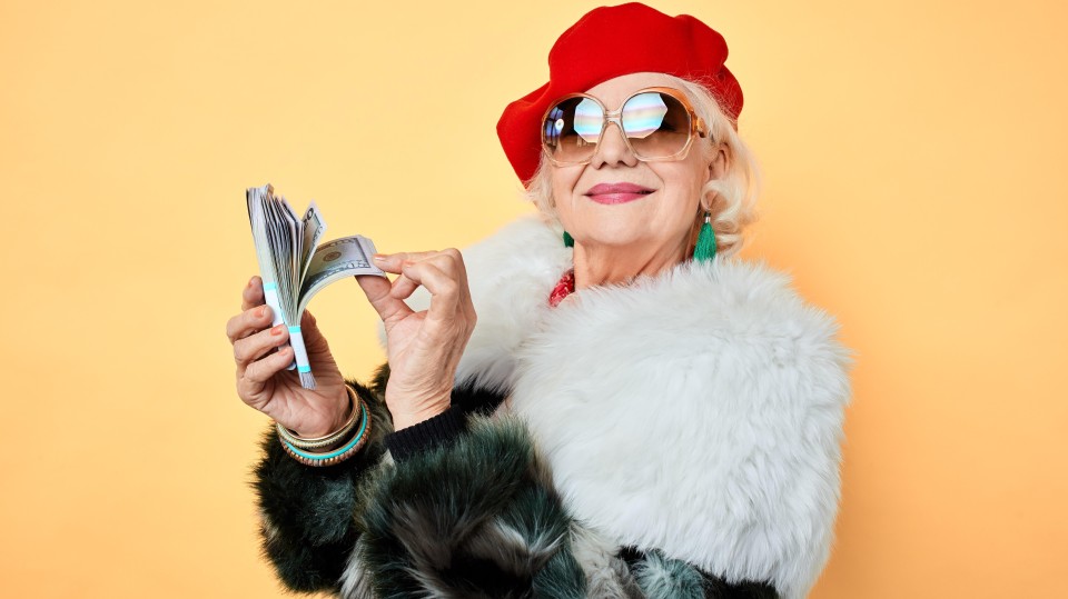 Een oudere vrouw in een bontjas, met een rode baret en met een grote zonnebril op, heeft een stapeltje bankbiljetten in haar handen.