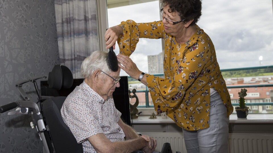 Vrouw met gele blouse kamt het haar van oudere man met witte blouse in rolstoel binnen.